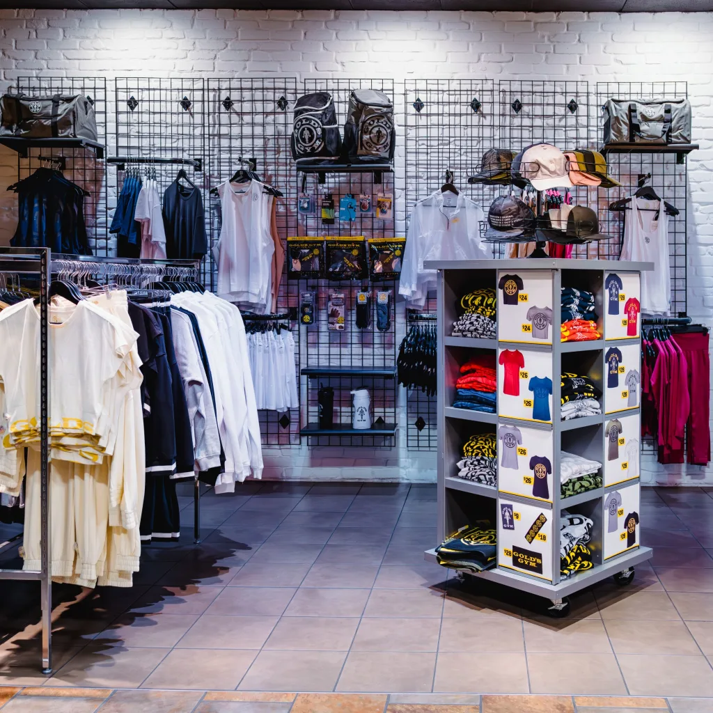Gym retail area with racks of clothing, hats, and accessories displayed against a white brick wall.