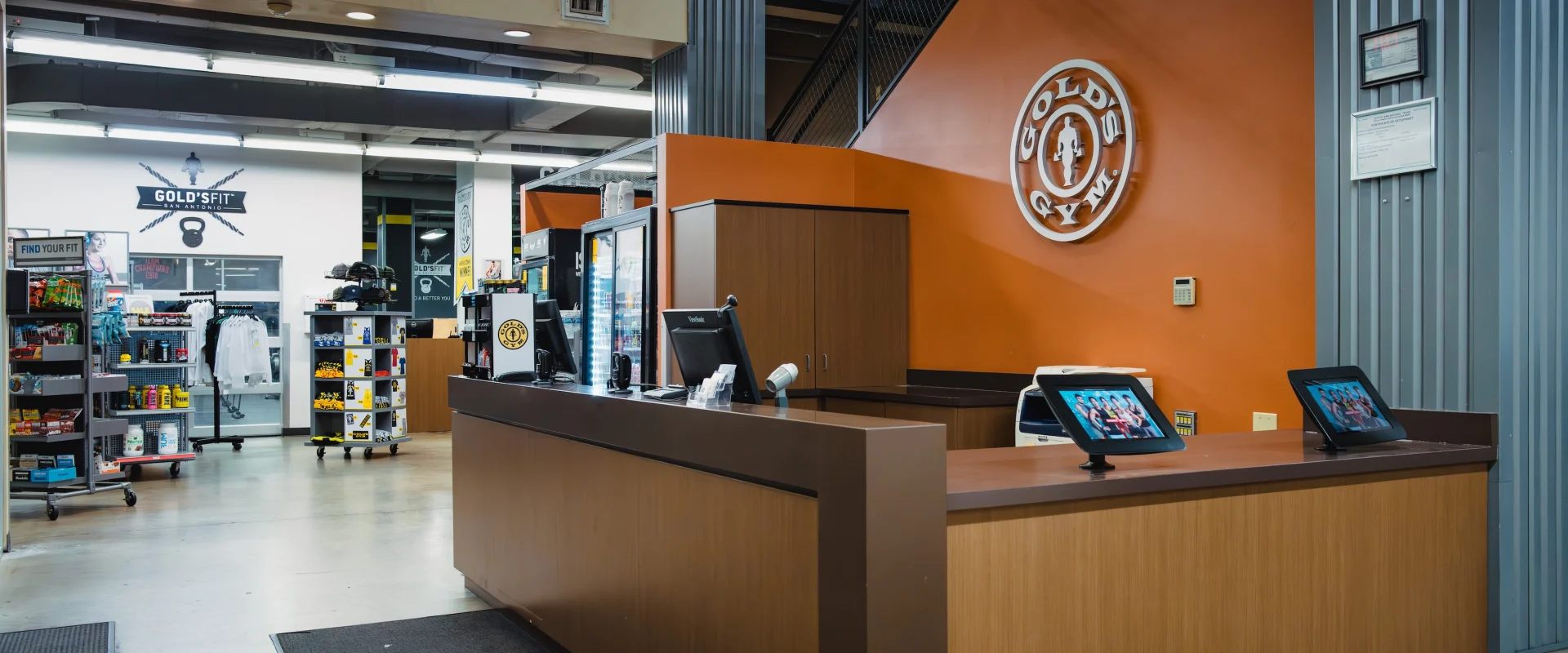 Gym reception area featuring a wooden desk with tablets and a logo on an orange wall. Shelves display snacks and merchandise, creating a welcoming atmosphere.