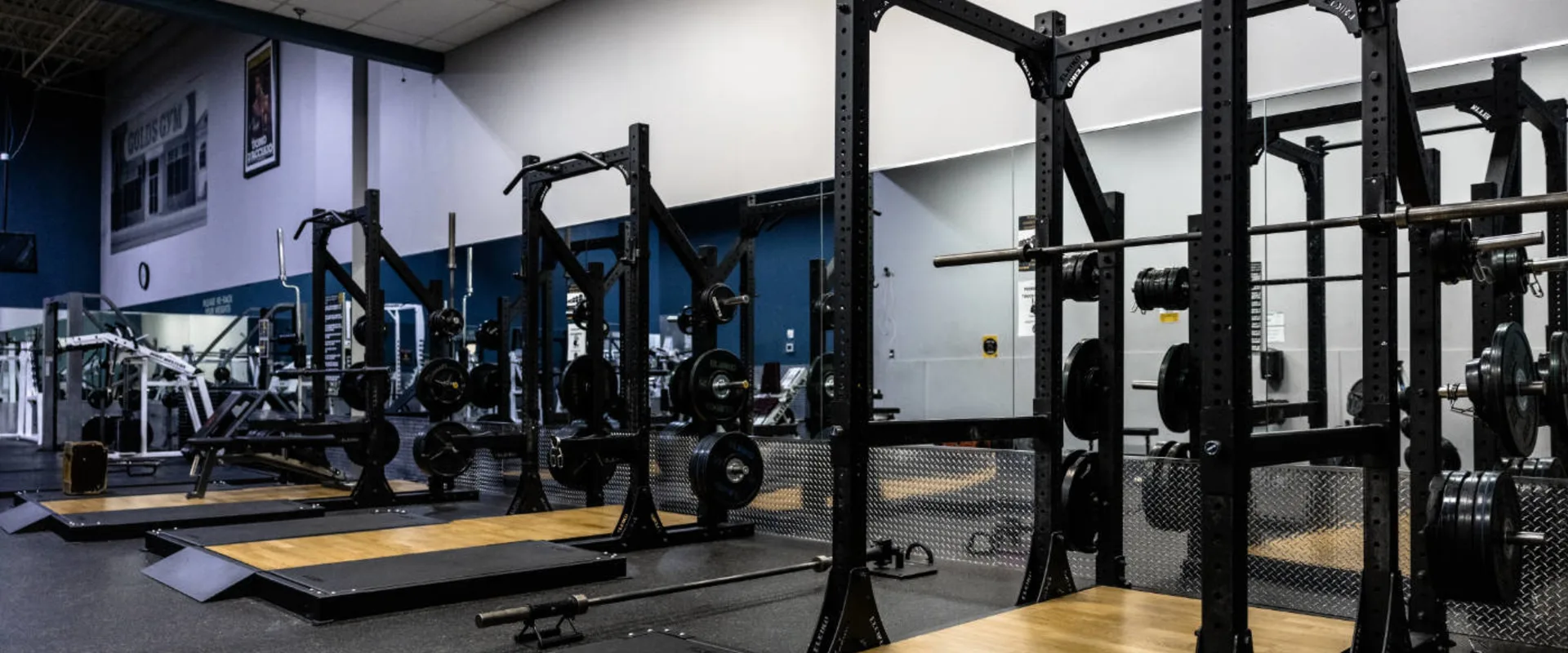Olympic lifting platforms at Gold’s Gym in Woodbridge (Lake Ridge), featuring black squat racks with barbells and weight plates. The platforms have a wood center with rubberized edges for shock absorption. Large mirrors line the wall, reflecting additional strength equipment in the background.