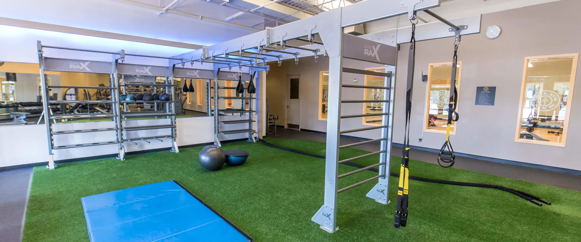 A spacious indoor gym with various exercise equipment and mats on a green artificial turf floor, surrounded by shelves and storage units.