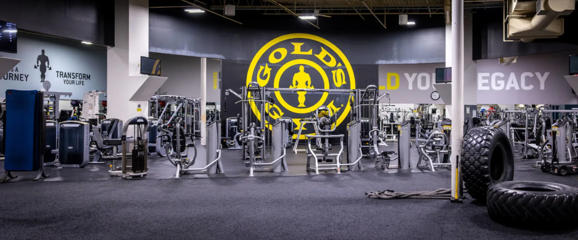 A variety of strength machines are arranged in rows beneath a large Gold’s Gym logo on the back wall. The area features selectorized equipment for resistance training. In the foreground, tractor tires and battle ropes sit on a turf section, contrasting with the rubber flooring. Mounted TVs line the walls, and mirrors reflect the equipment.