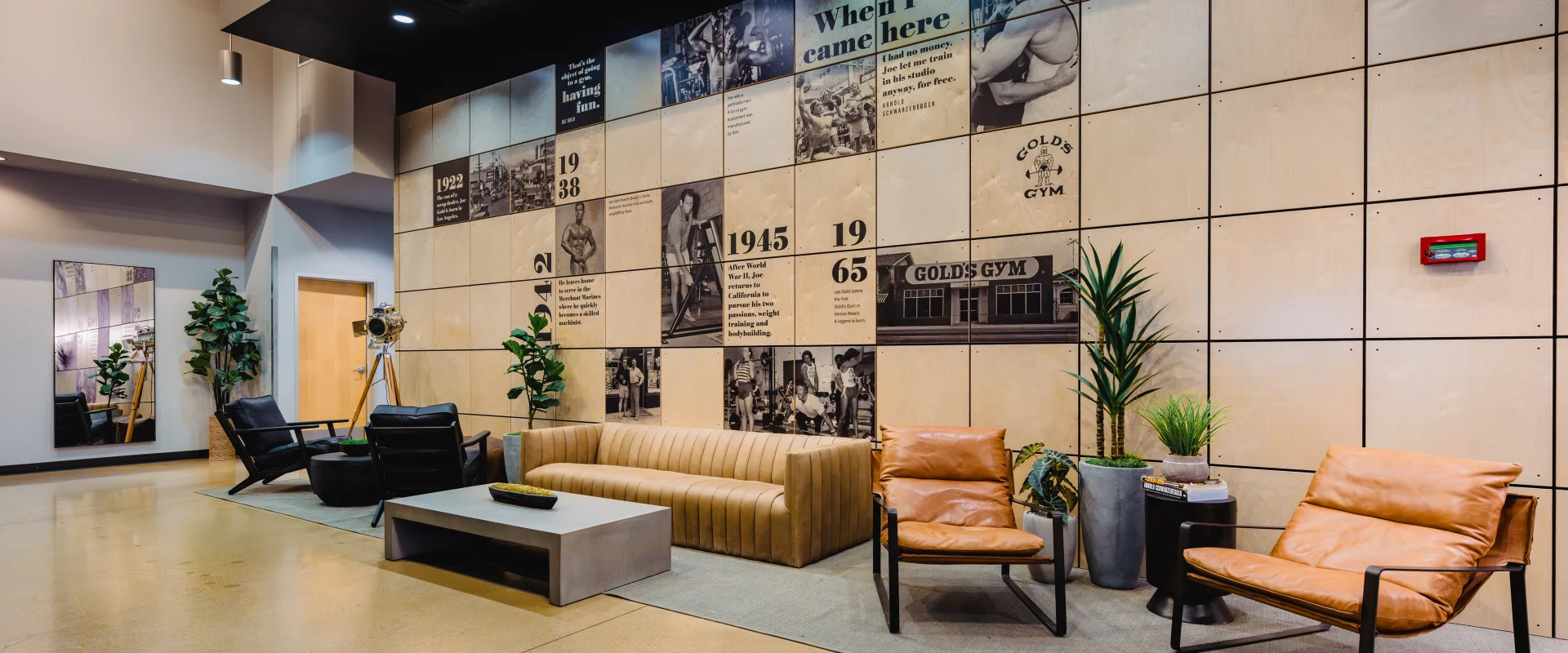 A cozy lounge area at Gold's Gym featuring a tan couch, leather chairs, modern decor, and a wall showcasing the gym's history with vintage photos and milestones.