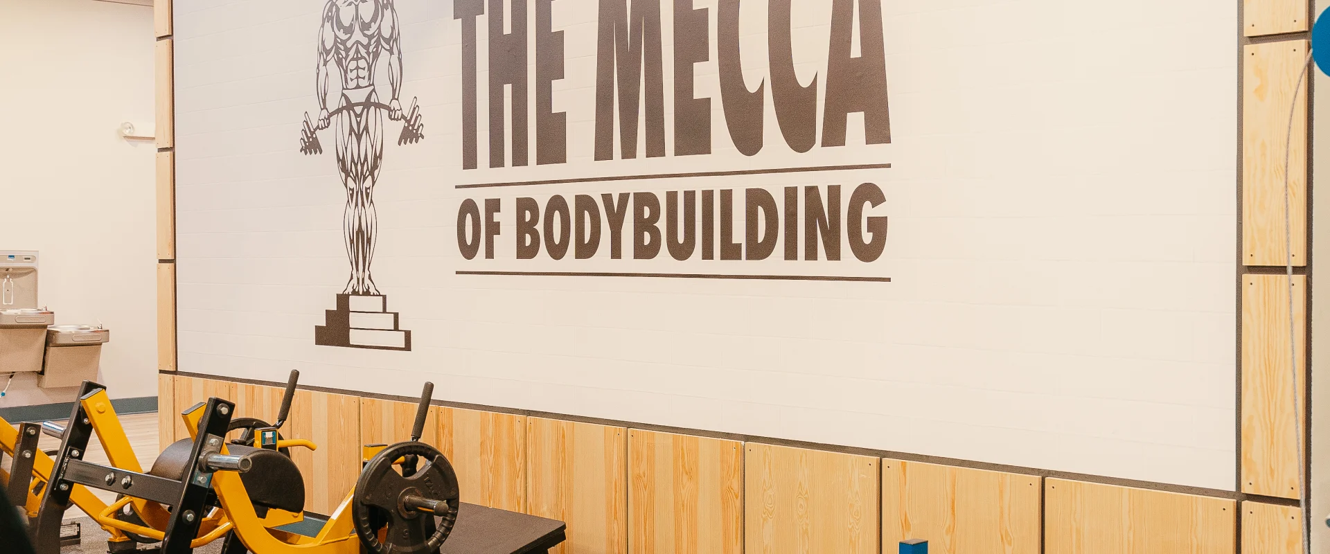 A wall in the free weights area at Gold's Gym Hendersonville, featuring a mural that reads "The Mecca of Bodybuilding" with an illustration of a muscular figure holding weights. The area also includes a hip thrust machine and a variety of other equipment.