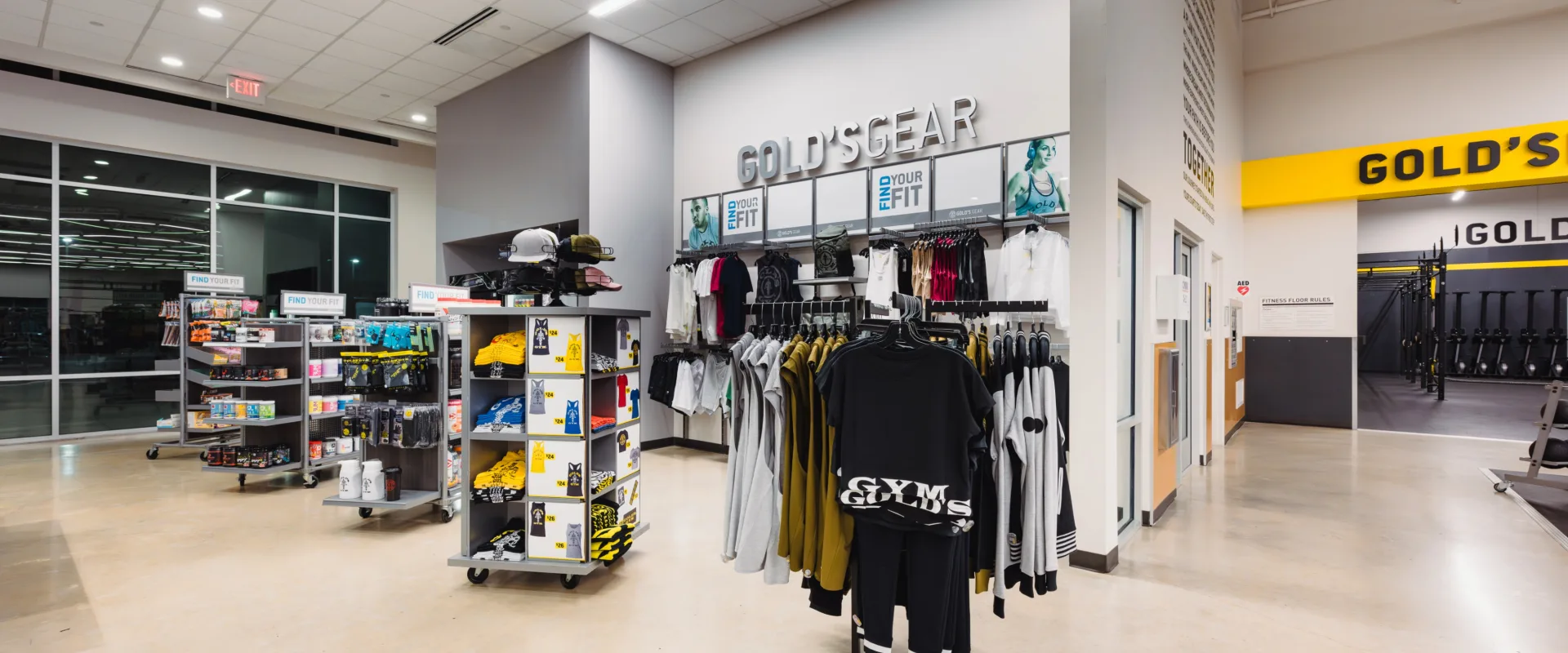 Pro Shop at Gold’s Gym in Texas (N. Round Rock), featuring a retail area with shelves stocked with fitness apparel, supplements, snacks, and accessories. The space includes racks of branded t-shirts, hoodies, and hats, along with neatly organized displays of water bottles, energy bars, and workout gear. Bright overhead lighting illuminates the open layout, and large windows allow natural light to enter the space. The "GOLD'S GEAR" signage is prominently displayed above the merchandise, and the adjacent gym area is visible in the background.