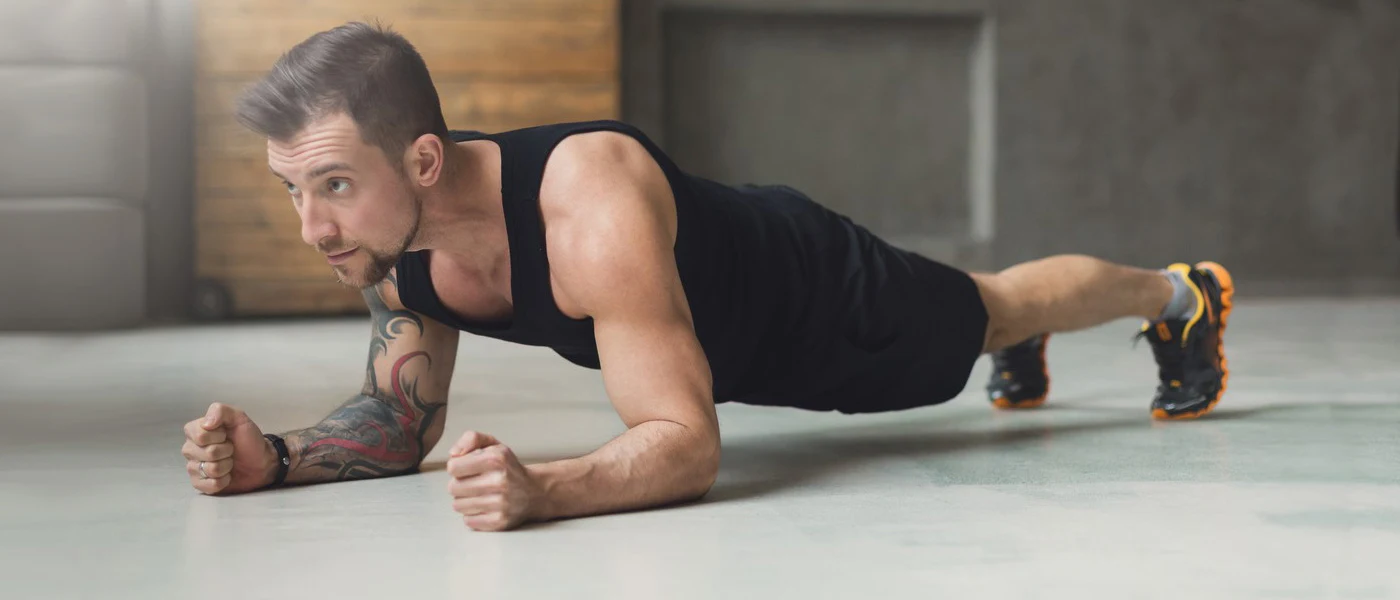 A person in a black tank top and shorts is performing a plank exercise on a gray floor, with a wooden wall visible in the background.