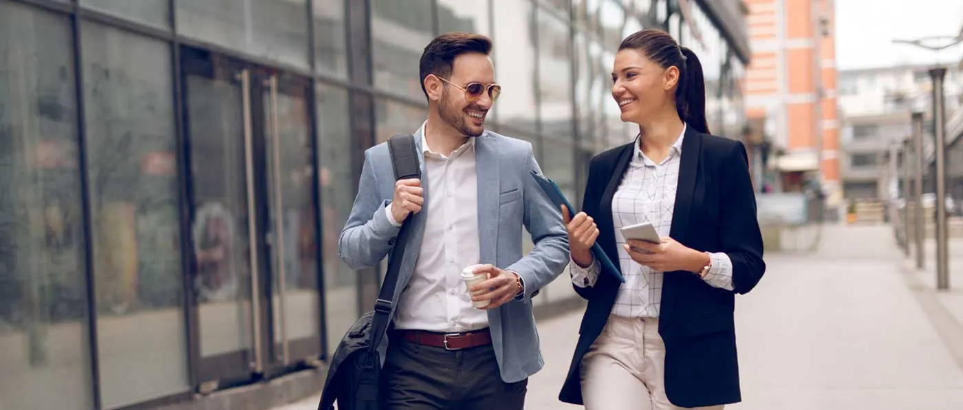 Two well-dressed individuals, a man and a woman, are engaged in a friendly conversation while walking along a city street with glass-paneled buildings in the background.