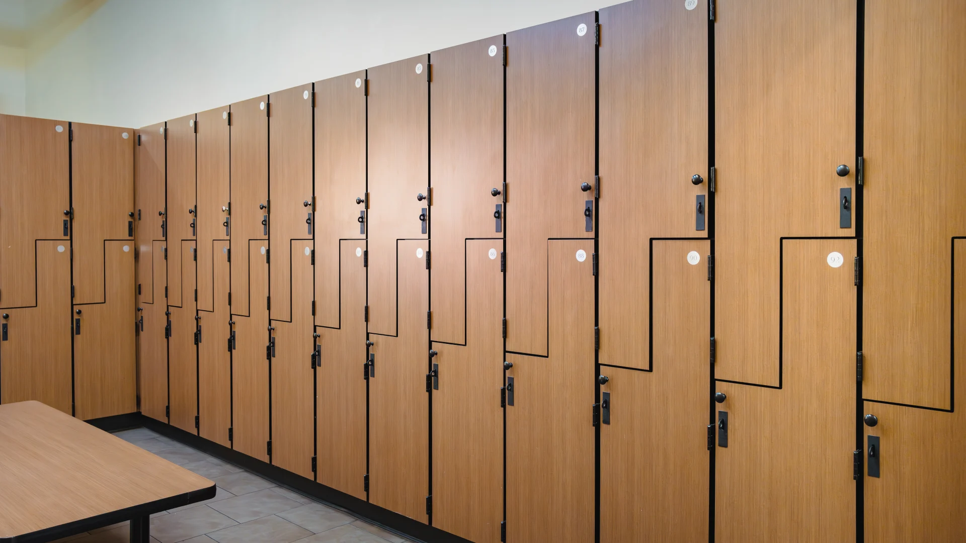 Locker room at Gold's Gym Austin (Downtown) featuring rows of wood-grain lockers with black hardware. A bench with a matching wood finish sits in front of the lockers, and the floor is tiled with light-colored ceramic.