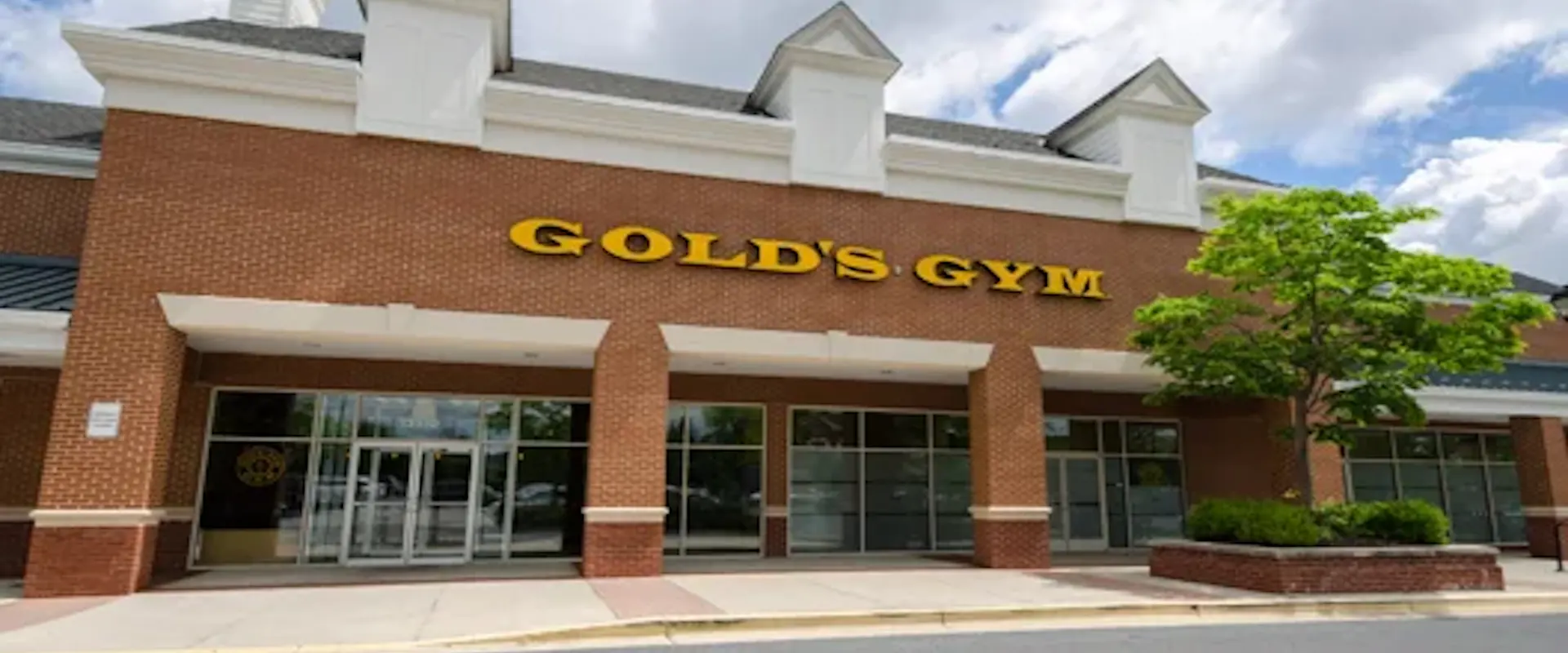 Exterior of Gold’s Gym Bowie with main entrance, brick facade, and yellow logo sign on a sunny day.