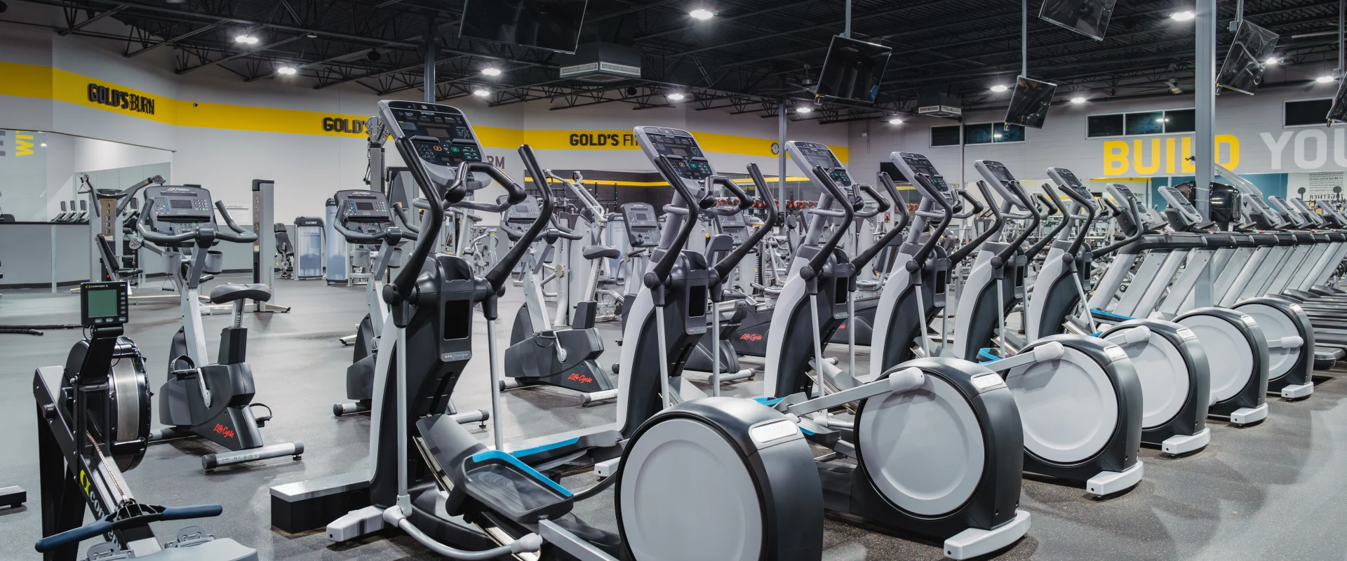 Row of elliptical machines and stationary bikes at Gold's Gym Belterra, arranged on a dark rubberized floor under bright lighting. Large overhead TVs and motivational signage, including "Build Your Body," are visible in the background, creating an inspiring workout space.