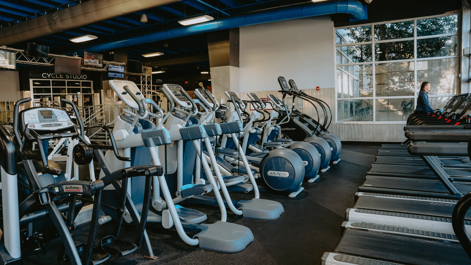 Cardio area at Gold’s Gym Tennessee, Downtown Chattanooga, featuring rows of elliptical machines, treadmills, and an Arc Trainer on a dark rubber floor. The space includes a high ceiling with exposed blue ducts, overhead lighting, and large windows showcasing natural light. A Cycle Studio entrance and mounted televisions are visible in the background.