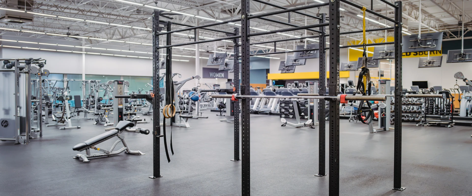 Functional training area at Gold's Gym in Texas (Pflugerville), featuring a versatile rig with pull-up bars, suspension trainers, gymnastic rings, and barbell attachments. The space is surrounded by various strength and cardio equipment, providing an open layout for dynamic workouts under bright, high-ceiling lighting.