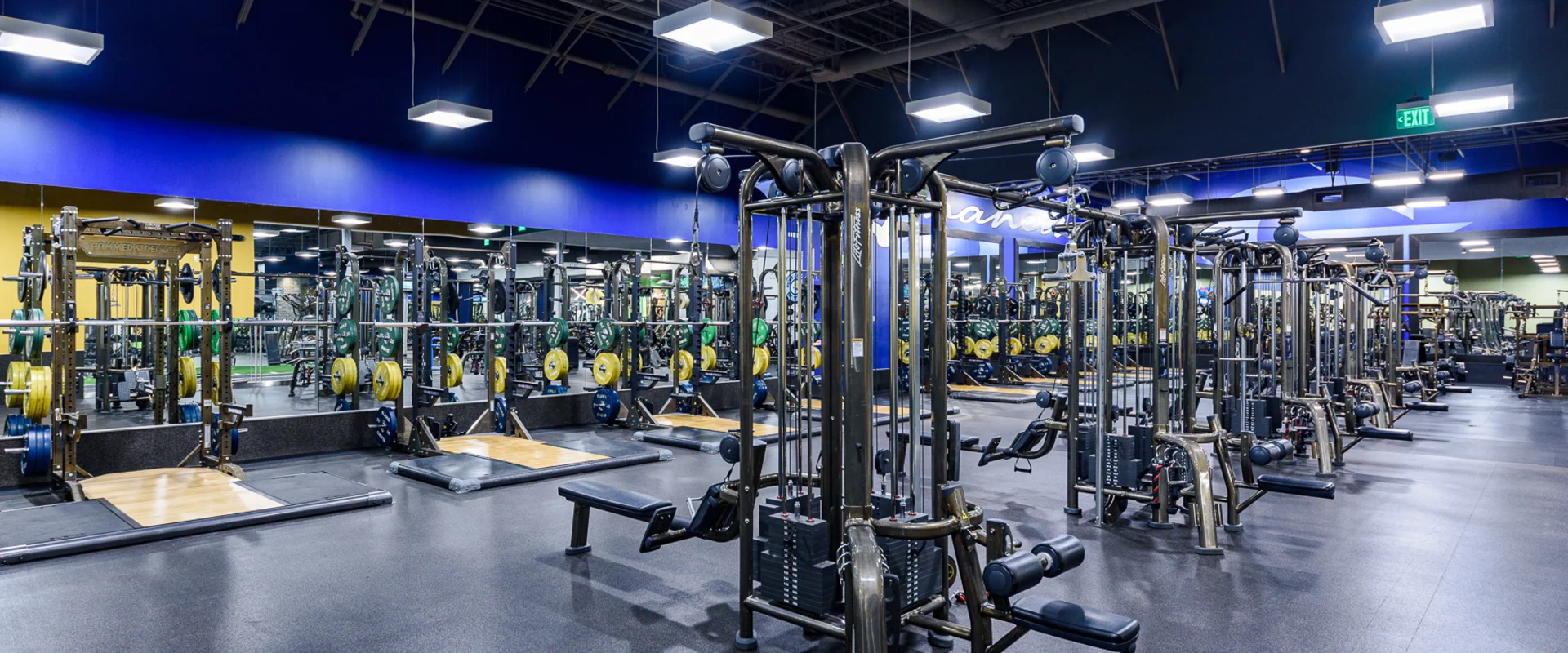 Machine weight area with cable machines, squat racks, and Olympic lifting platforms. Mirrors line the walls, reflecting rows of weightlifting equipment. Overhead lighting highlights the spacious, clean gym layout.