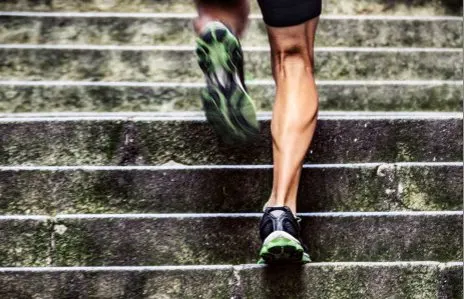 The image shows a person's legs and feet wearing athletic shoes with green and black patterns, walking up a set of concrete stairs against a blurred background.