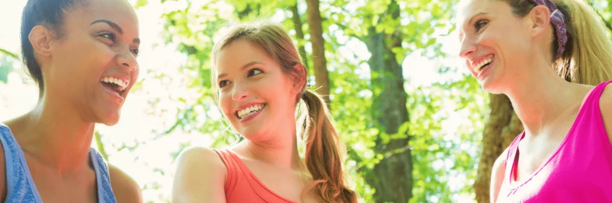 Three smiling people, two women and one man, are captured in a lush, green outdoor setting with trees and foliage in the background.