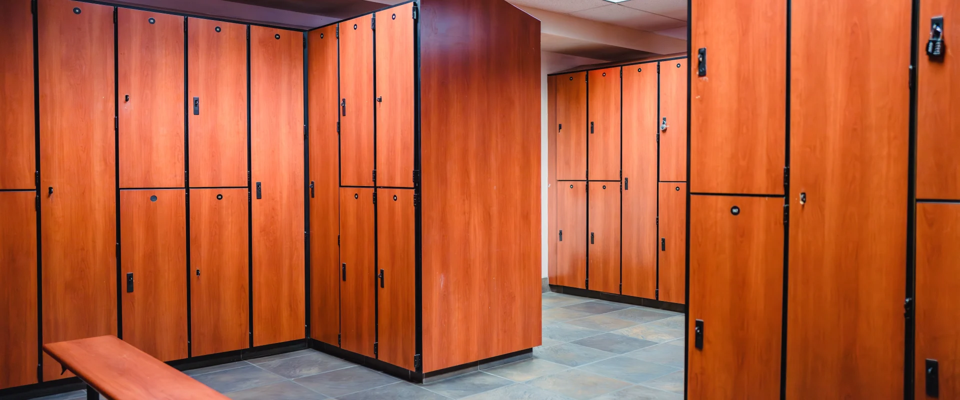 The locker room at Gold’s Gym in Austin (South) features rows of tall, wood-finished lockers with black trim, creating a warm and clean aesthetic. Overhead fluorescent lighting brightens the space, reflecting off the sleek, tiled floor. A simple wooden bench with metal legs sits in the center, providing seating for convenience. The layout is spacious, with wide walkways for easy access to lockers, enhancing both functionality and comfort.