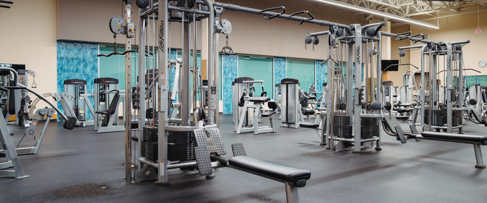 Spacious gym with various exercise machines, including weight stacks and benches, under bright fluorescent lights. The space is clean and organized.
