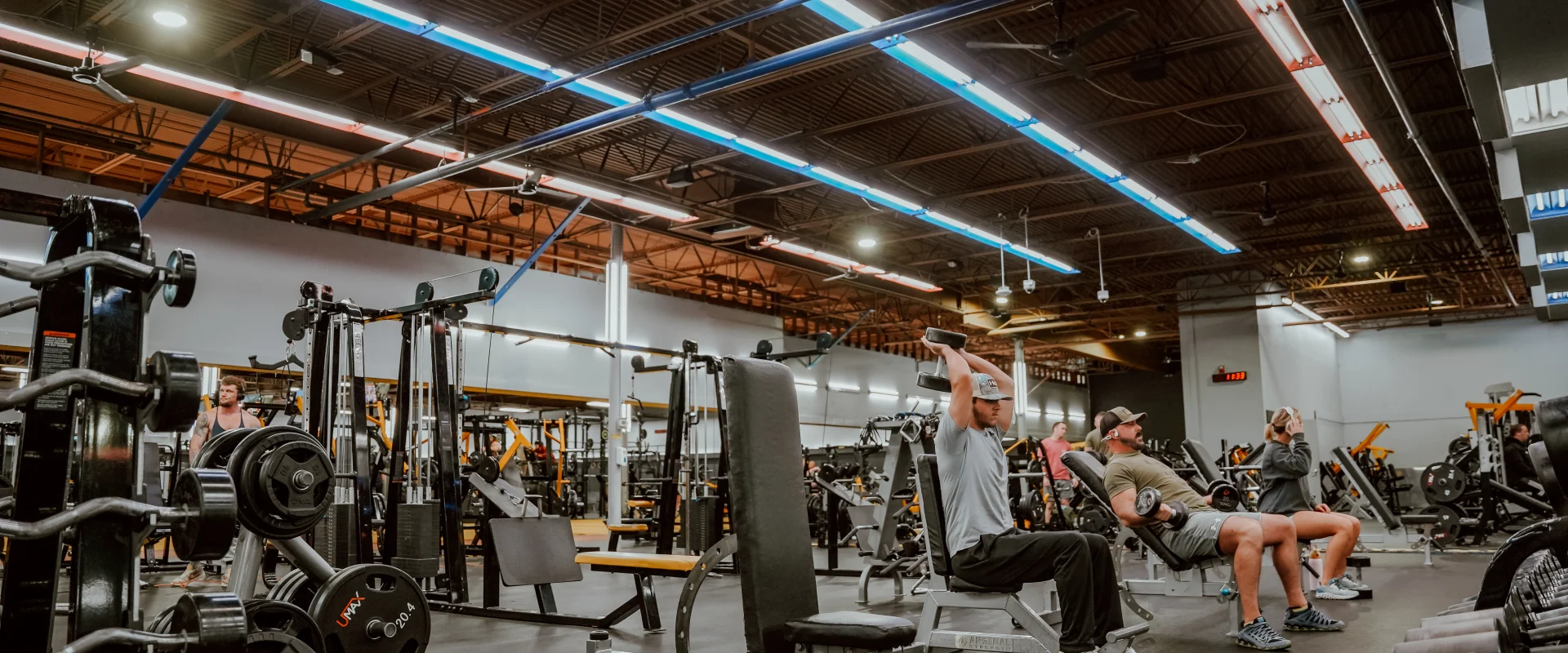 Dumbbell area at Gold's Gym Murfreesboro North, showing gym-goers using benches and free weights under industrial-style ceilings with bright overhead lighting. Rows of dumbbells and weight plates are visible in the foreground.