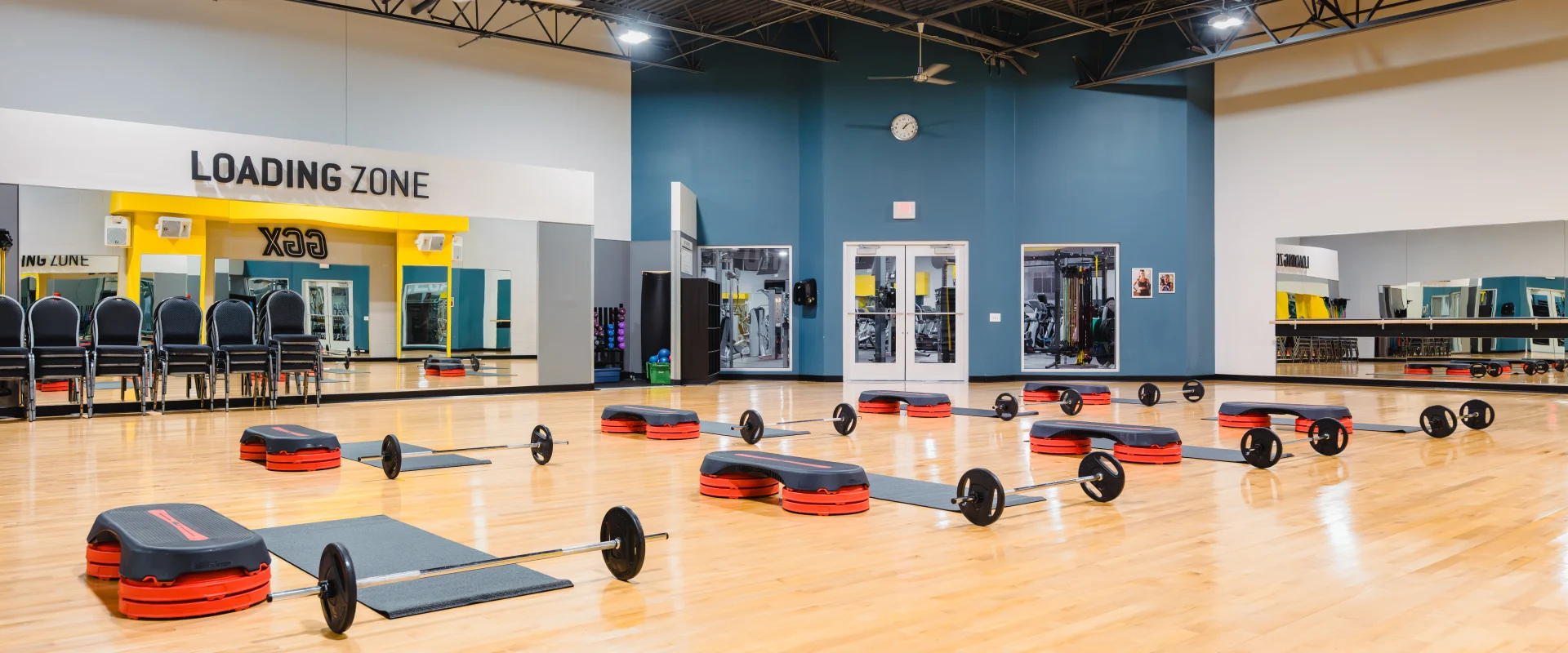 Group exercise studio with red and black step platforms, barbell sets, and mats arranged on a polished wood floor, surrounded by mirrors and blue walls under a high ceiling.