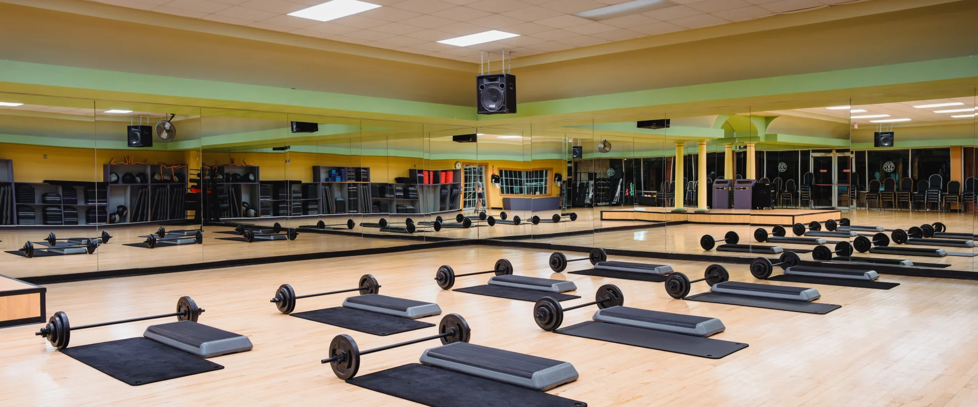 Spacious gym room with a mirrored wall, wooden floor, and ceiling lights. Barbells on mats are neatly arranged, creating a calm and organized atmosphere.