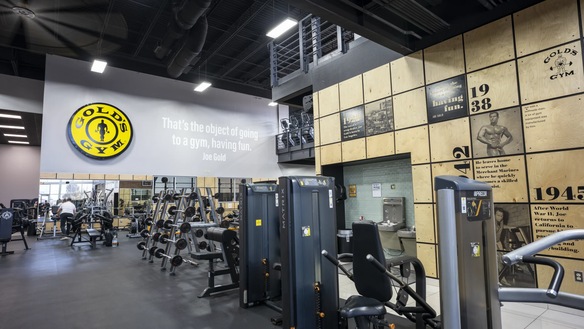 An interior view of a Gold’s Gym weight room. A large gray wall features a prominent yellow and black circular logo and a quote by Joe Gold: "That's the object of going to a gym, having fun." To the right, a multi-paneled wood wall displays historical timelines and photos of the gym's heritage. In the foreground, various black selectorized strength machines and dumbbell racks are arranged on a dark gym floor beneath a black industrial-style ceiling.