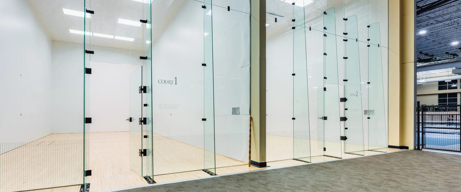 Bright, modern indoor racquetball court with floor-to-ceiling glass walls and a carpeted exterior hallway, conveying a clean, sleek, and open atmosphere.