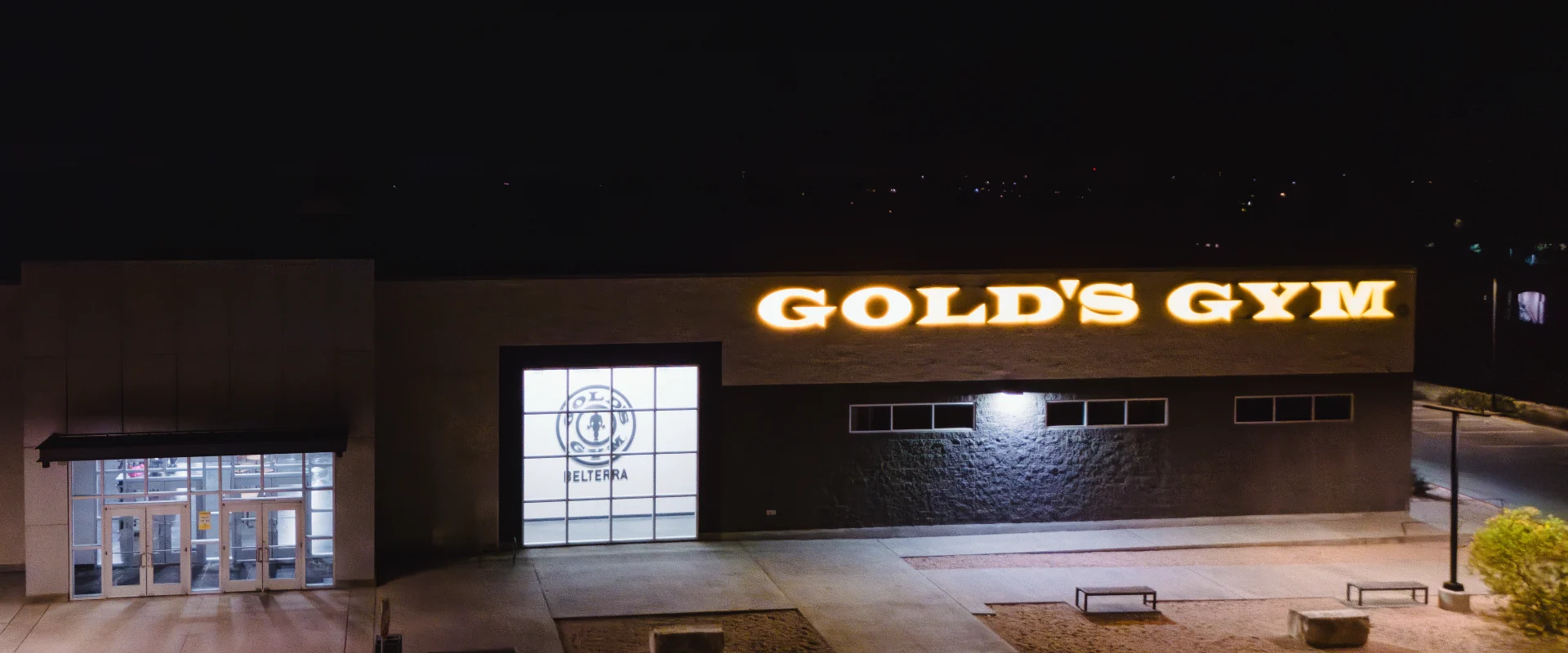 Exterior of Gold's Gym Belterra at night, showcasing the illuminated Gold's Gym signage on the building facade. The entrance features large glass doors, and the gym's logo is visible through a well-lit window. The surrounding area includes paved walkways, benches, and minimal landscaping.