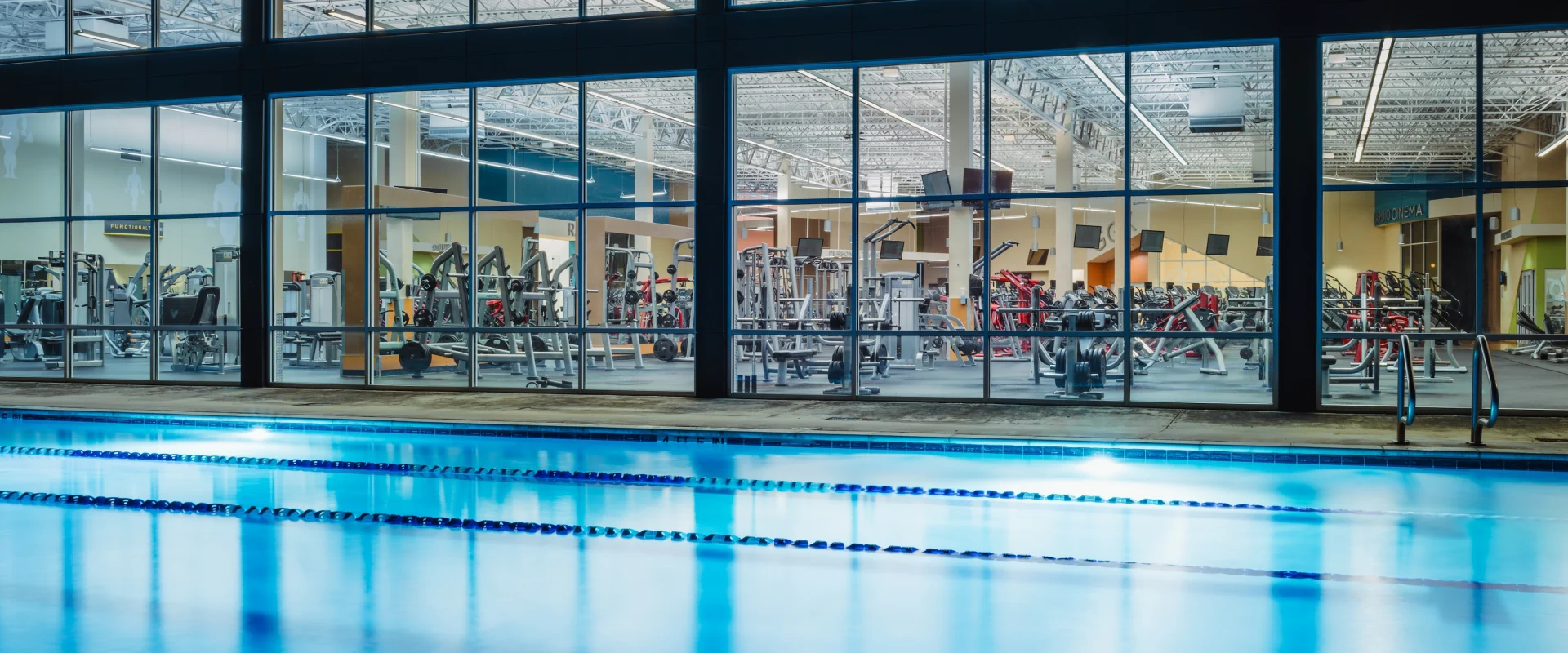 View of a gym through large windows, showcasing various workout equipment. In the foreground, a tranquil indoor pool reflects blue lighting.