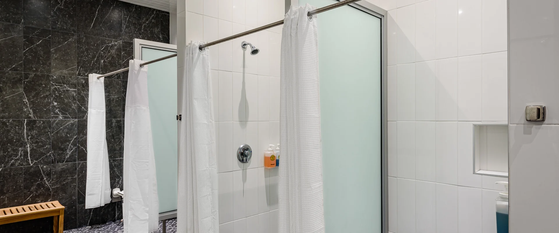 The shower area at Gold’s Gym Cerritos features private shower stalls with frosted glass doors and white curtains. The walls are covered in glossy white tiles, while the floor has a black and gray mosaic tile pattern. A wooden bench sits against a black marble accent wall. Overhead recessed lighting illuminates the space, and built-in soap dispensers are mounted inside the showers.