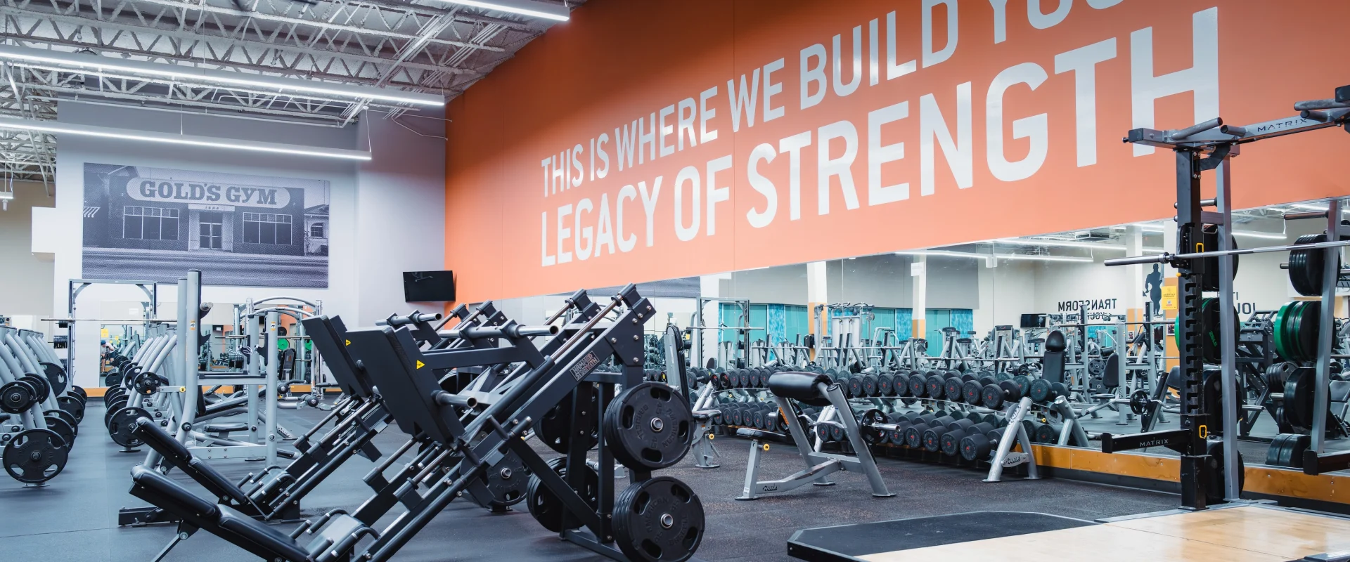 Modern gym interior with weightlifting equipment, a bold orange wall reading "This is where we build your legacy of strength," and motivational ambiance.