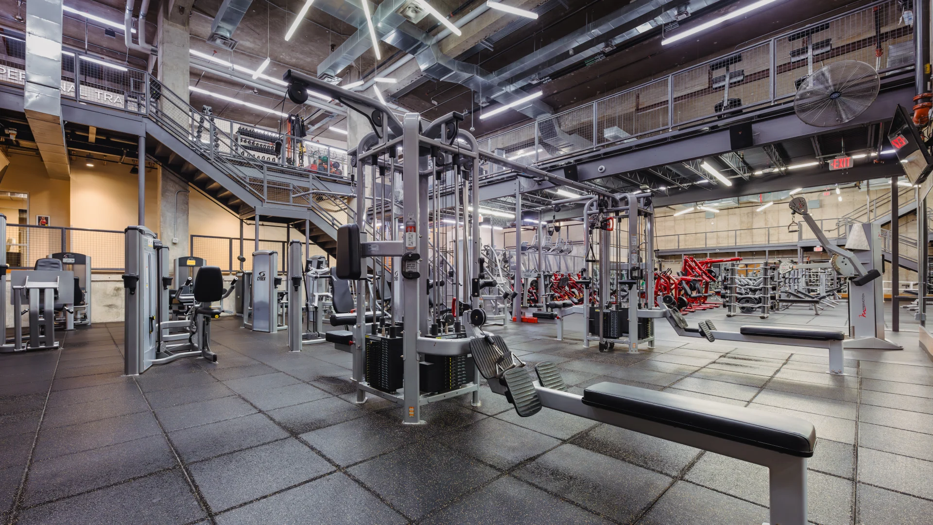Machine weight area at Gold's Gym Austin (Downtown) featuring strength training equipment on a black rubber floor, with industrial-style lighting and exposed ceilings. A mezzanine staircase leading to an upper-level workout area visible in the background.