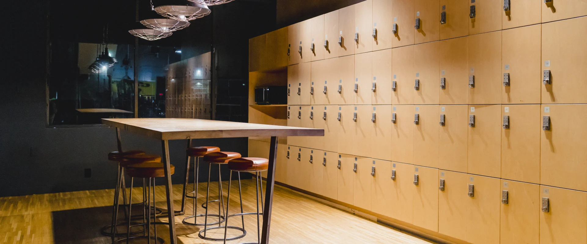 Dimly lit locker room area featuring wooden floors, lockers with free locks, a wooden table with metal legs, empty leather stools, and large softly illuminated light fixtures above the table