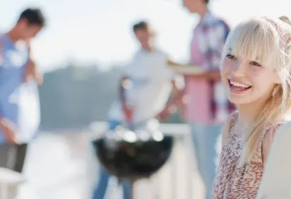 A smiling young woman in the foreground, with a group of people in the background, appears to be at some kind of social gathering or event.