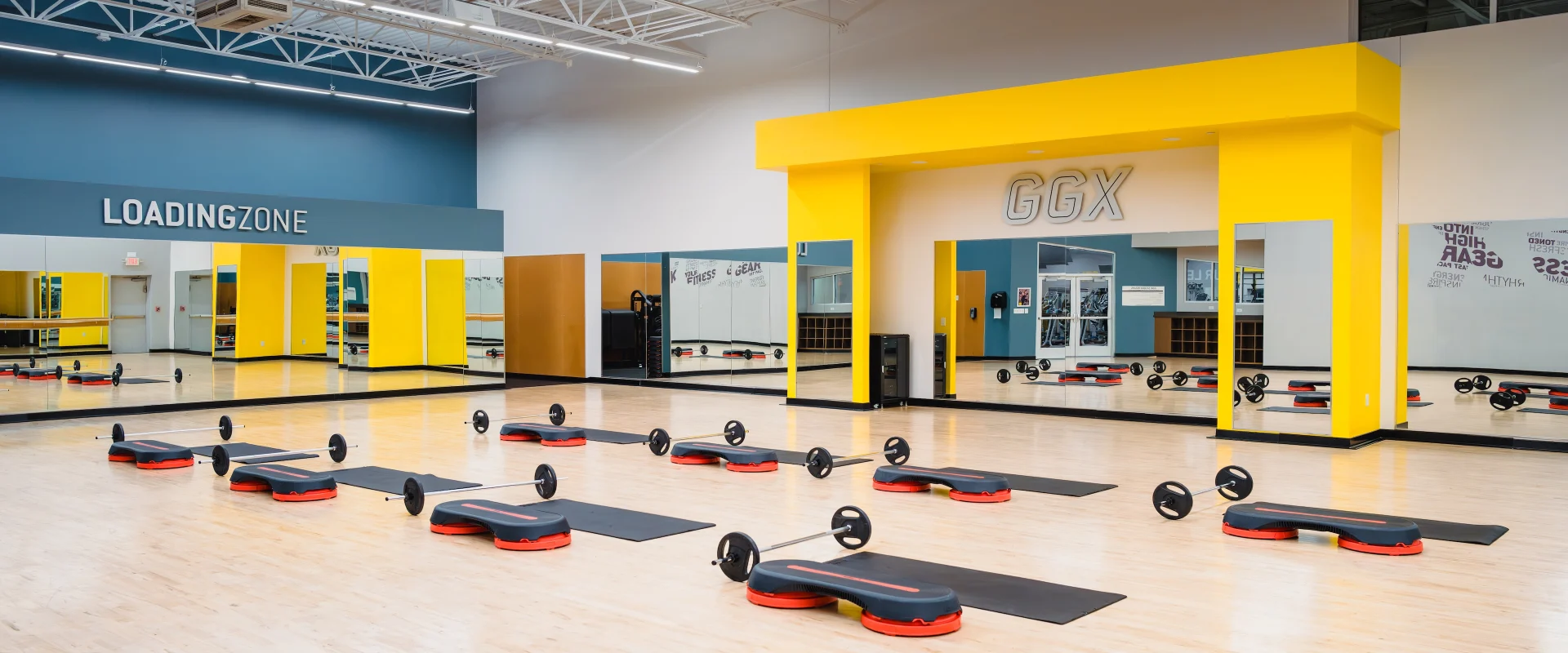 Group Exercise studio at Gold’s Gym in Texas (N. Round Rock), featuring a spacious area with a polished wooden floor and large wall mirrors. The studio is set up with workout stations, including barbells, weight plates, aerobic steps with risers, and exercise mats arranged in rows. A bold yellow accent structure frames the front of the room with "GGX" displayed above. The mirrored walls reflect the equipment and create an open, bright environment. The space also includes a "Loading Zone" area with additional mirrors and equipment storage.
