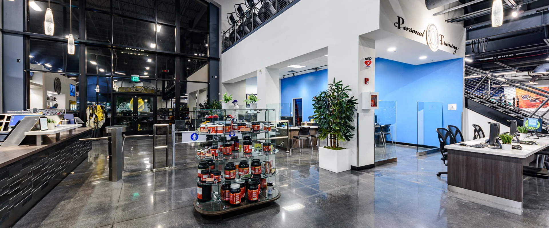 The gym’s front desk area features a sleek modern design with a polished concrete floor. A supplement display stands near the entrance, while personal training offices and gym access gates are visible in the background.