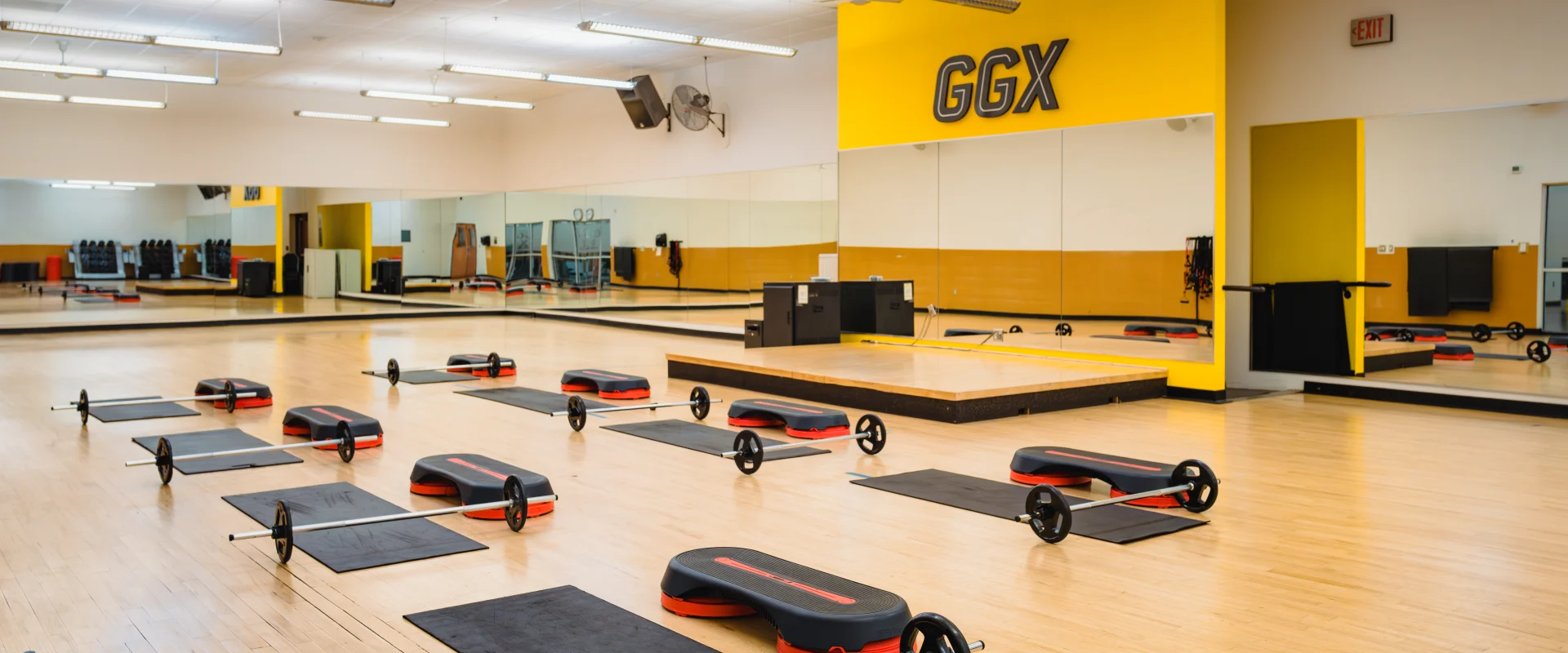 Group exercise room at Gold's Gym in Austin (Hesters Crossing) featuring wood flooring, mirrored walls, and yellow accents. Equipment, including barbells, mats, and steps, is evenly arranged for a class setup. A raised platform at the front serves as an instructor stage. The room is well-lit with overhead lights and ceiling fans.
