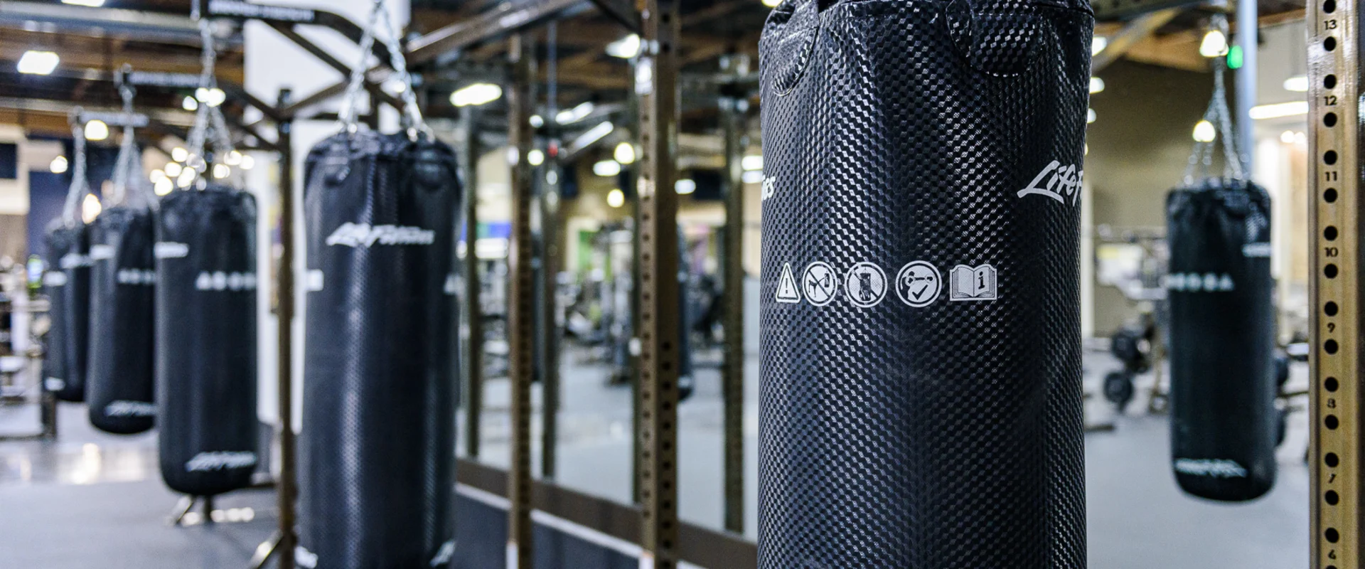 The boxing area at Gold’s Gym Cerritos features multiple black heavy bags hanging from a sturdy metal frame. Each bag is secured with metal chains. A mirrored wall runs along the space, reflecting the equipment and gym environment.