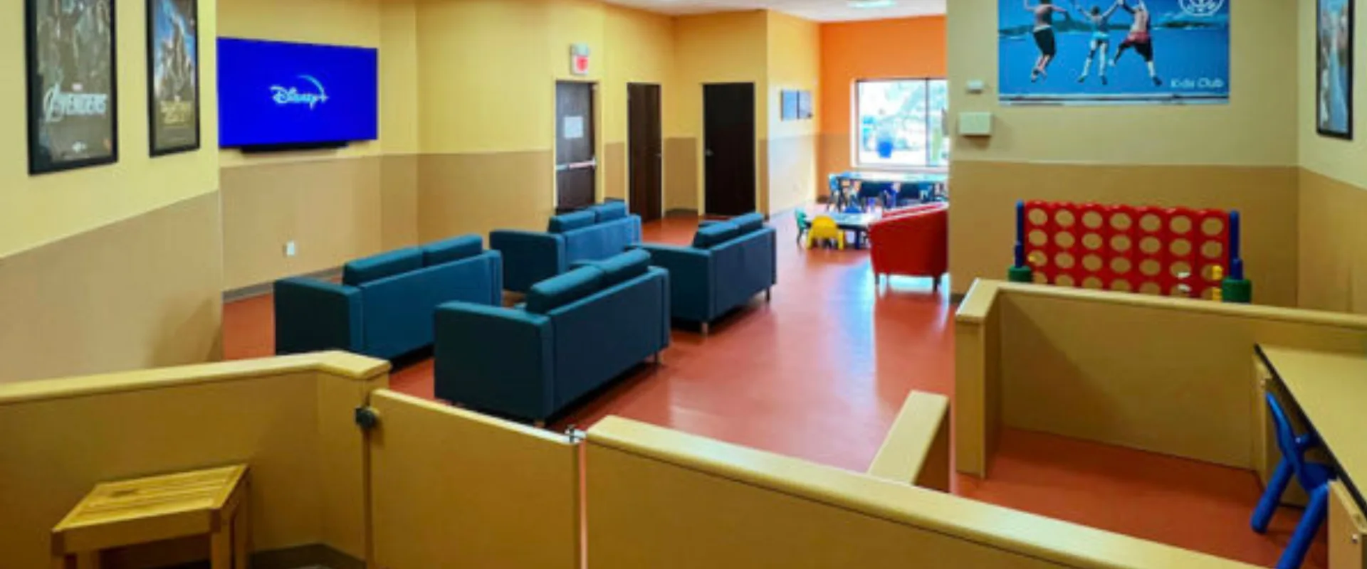 Kids Club at Gold’s Gym in East Northport, New York, featuring a play area with child-sized tables, colorful chairs, and a large Connect Four game. The room includes a seating area with blue armchairs facing a wall-mounted TV displaying Disney+, bright overhead lighting, and vibrant wall posters. Large windows provide natural light, creating a welcoming space for children.