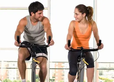 Two people, a man and a woman, are exercising on stationary bicycles in what appears to be a fitness studio or gym setting, with large windows visible in the background.