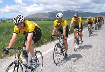 A group of cyclists in yellow jerseys riding on a road surrounded by mountains and fields under a cloudy sky.