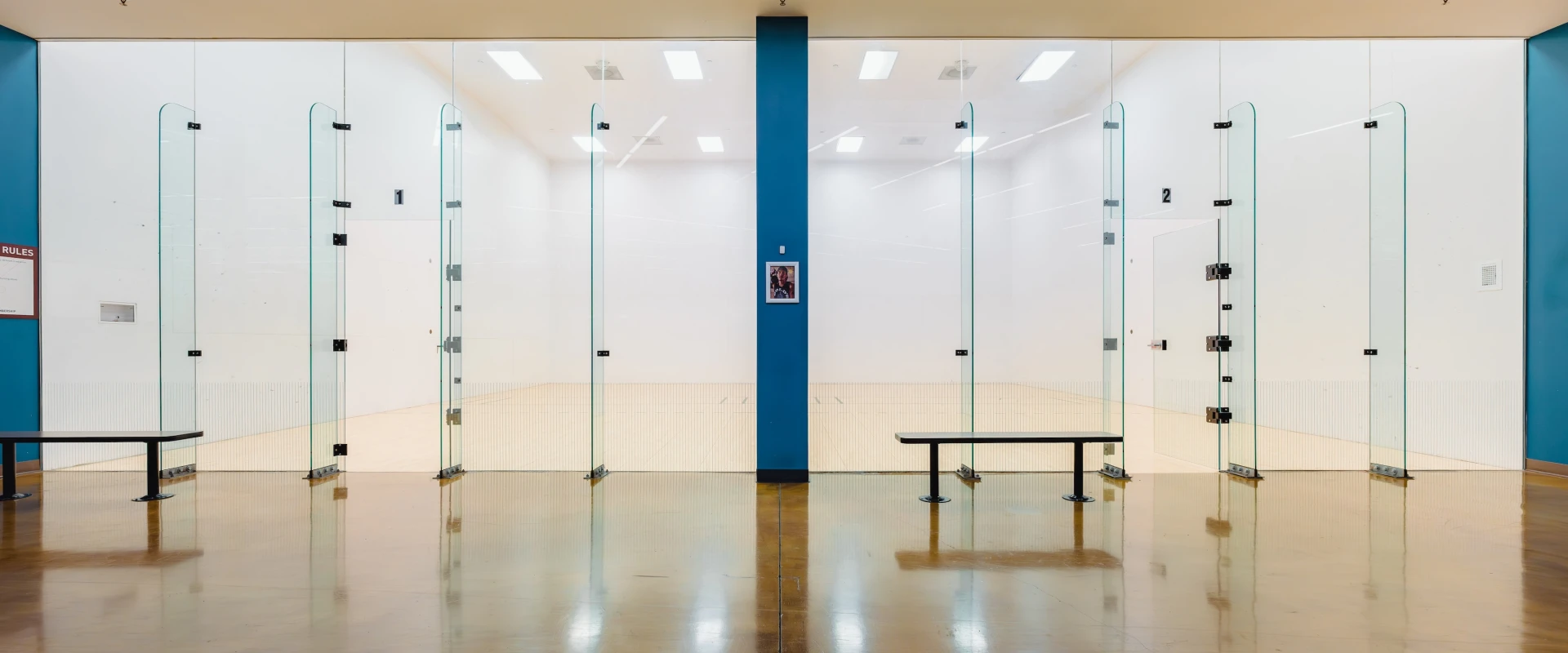 Well-lit racquetball court with glass walls, bench outside, and a bold "RACQUETBALL" sign above. Clean, modern, and ready for play.