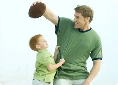 A man in a green shirt is holding a football and interacting with a young boy, both wearing similar green shirts, against a plain white background.