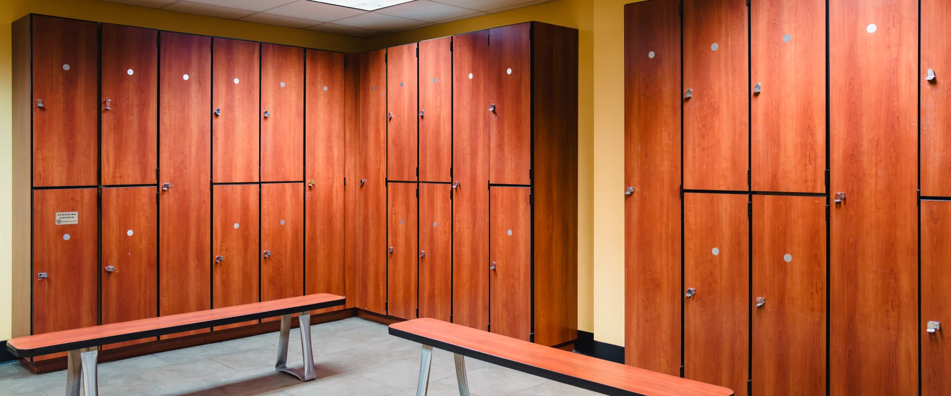 Gold's Gym locker room in Austin (Bee Caves) featuring rows of wooden lockers against a light yellow wall, tiled flooring, and matching wooden benches with metal legs in a well-lit space.