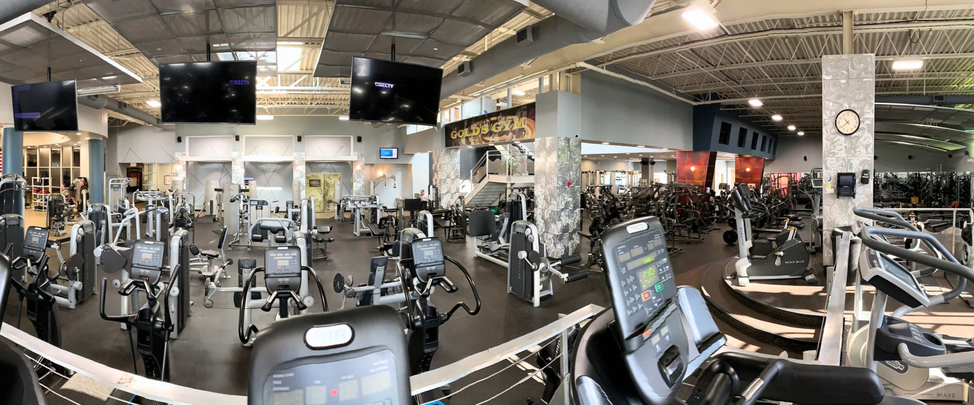 Resistance machine area at Gold's Gym in Smithtown, featuring rows of seated and standing strength training machines with digital displays. The foreground shows elliptical machines and stationary bikes with control panels, while the background includes strength machines organized into sections. Large flat-screen TVs hang from the ceiling, and the Gold's Gym logo is displayed above a staircase leading to a mezzanine. Bright lighting and polished flooring create a modern, organized atmosphere.