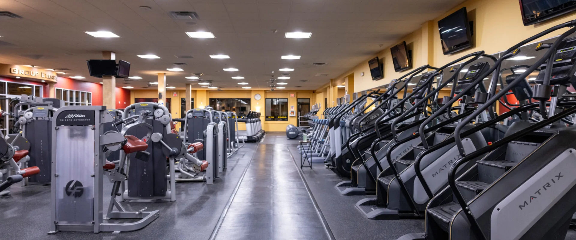 Strength equipment area at Gold’s Gym in East Northport, New York, featuring Life Fitness selectorized machines for targeted muscle training, including triceps extension and leg press machines. The space includes Matrix stair climbers and treadmills lined along mirrored walls with overhead TVs. Bright ceiling lights and rubber flooring create a spacious, organized workout environment.