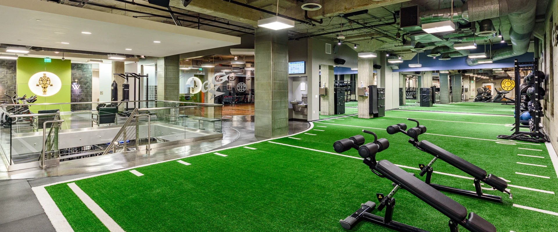 Turf area at Gold’s Gym Hollywood, featuring a large green artificial turf section marked with white lines for functional training. Two adjustable decline benches are positioned in the foreground. In the background, strength training equipment, punching bags, and weight racks are visible. A staircase with a glass railing leads to a lower level. Exposed ductwork, concrete pillars, and industrial lighting fixtures line the ceiling. A Gold’s Gym logo is displayed on a green accent wall near the boxing area.