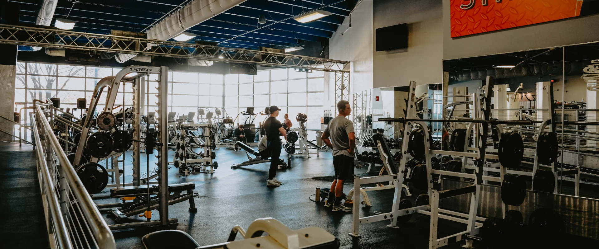 Free weights area at Gold’s Gym Tennessee, Downtown Chattanooga, showcasing weight benches, racks of weight plates, and a Smith machine on a dark rubber floor. The space features large windows allowing natural light, a high ceiling with exposed blue ducts, and overhead lighting. Gym members are visible utilizing the equipment, with additional workout areas and a "Strength" motivational banner in the background.