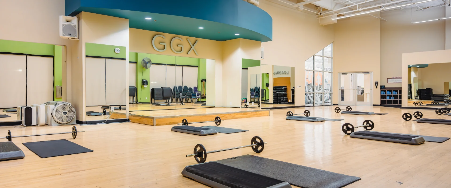 Group Exercise studio in Austin (Westlake), featuring a spacious hardwood floor setup with mats, barbells, and risers neatly arranged for class participants. The room includes a raised instructor stage under a bold blue arch labeled “GGX,” large mirrors along the walls, and ample natural light from floor-to-ceiling windows. The open, airy environment is perfect for dynamic group workouts.