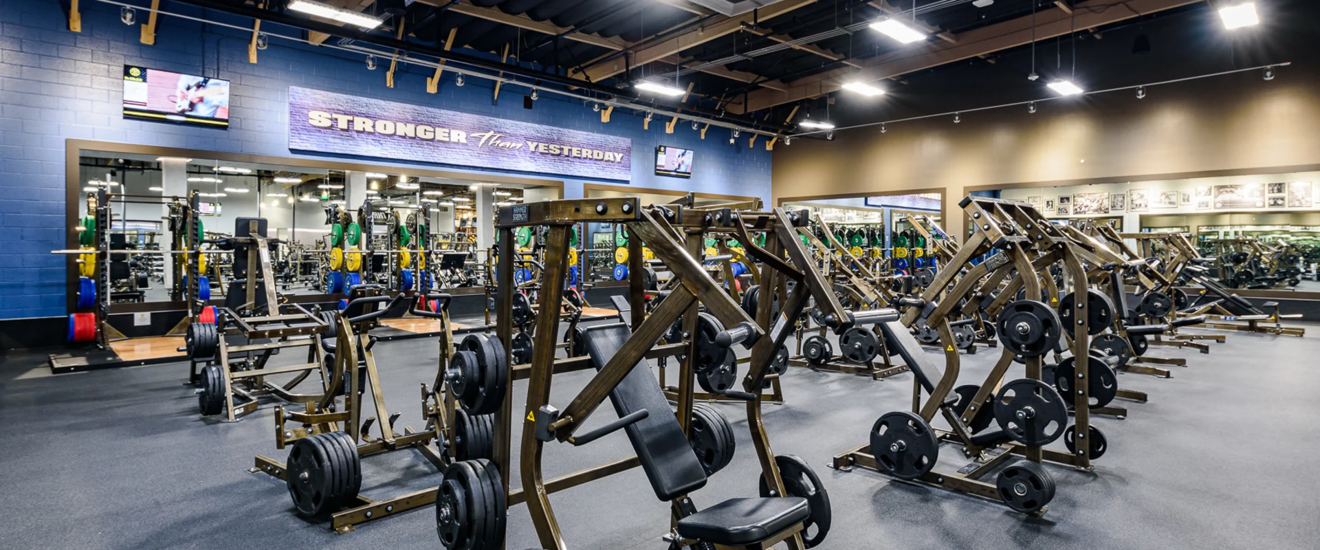 The weight machine area at Gold’s Gym Cerritos includes various strength machines with black padding and metallic frames, positioned on black rubber flooring. A mirrored wall reflects the space, and a ‘Stronger Than Yesterday’ sign is displayed above the equipment. Overhead lighting and exposed beams contribute to the open gym layout.