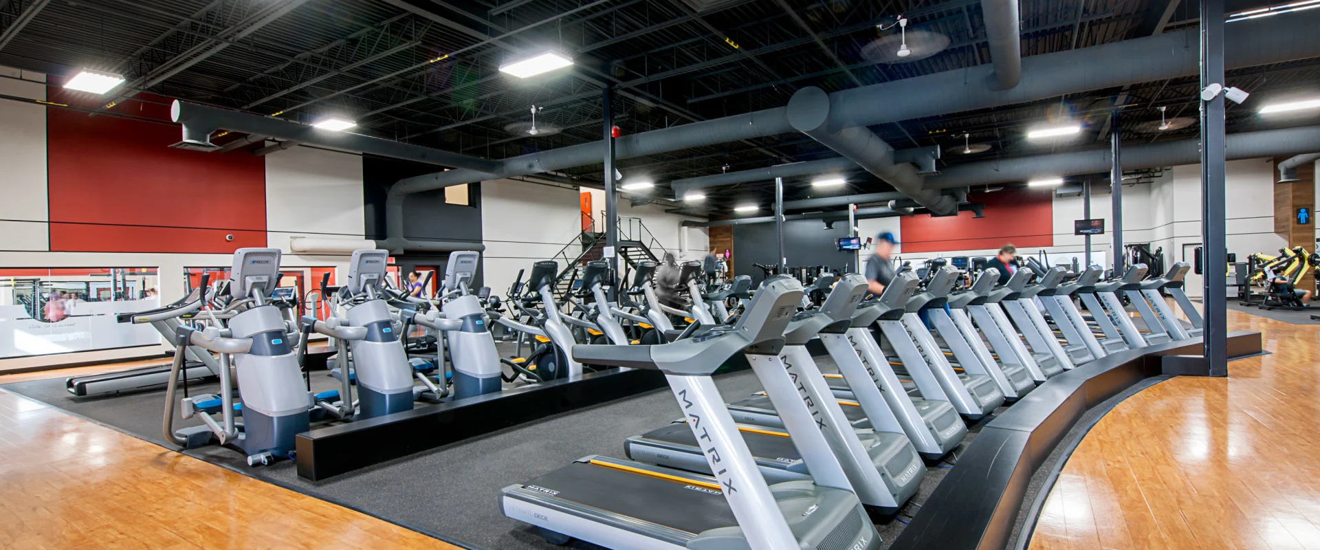 Cardio area at Gold's Gym Vertu featuring rows of Matrix treadmills and elliptical machines on raised black flooring, surrounded by warm-toned wood floors and overhead industrial lighting.