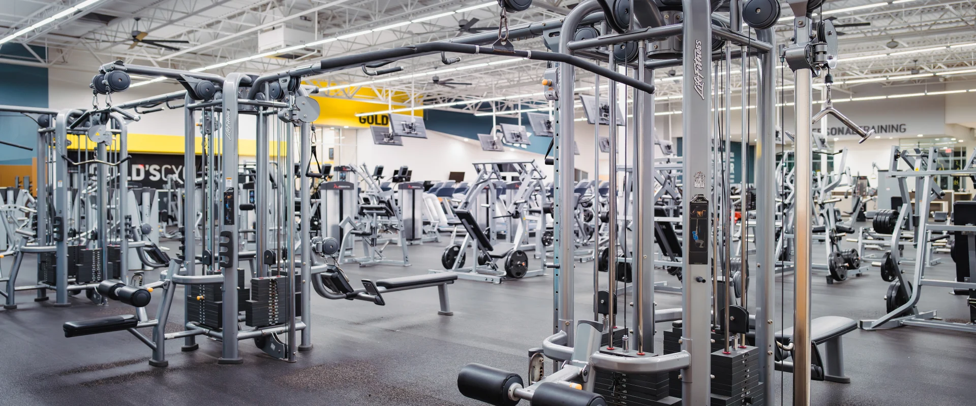 Resistance Machines at Gold's Gym in Texas (Pflugerville), featuring sleek, silver cable machines with adjustable weight stacks. The spacious layout includes benches, pulley systems, and a variety of strength training equipment, set against a backdrop of bright lighting and modern gym design.
